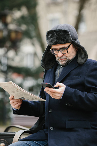 Un anciano disfruta de una fría mañana de invierno en la ciudad. Un hombre se sienta en un banco en la plaza, lee un periódico y charla alegremente con amigos en un teléfono móvil. - Foto, Imagen