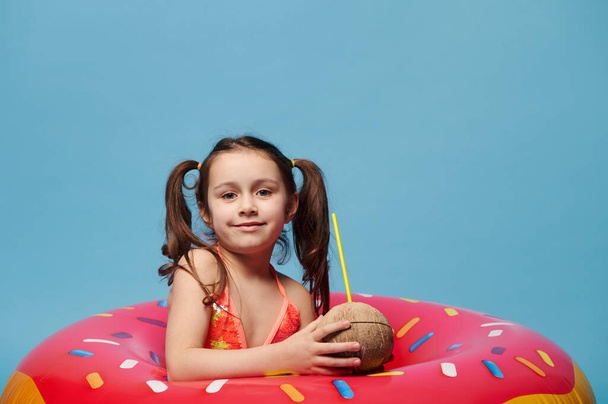 Υπέροχο κοριτσάκι με δροσιστικό coconut cocktail με καλαμάκι στα χέρια, χαμογελά έντονα κοιτάζοντας την κάμερα, ποζάροντας μέσα σε ένα φουσκωτό πολύχρωμο donut swim ring, σε φόντο μπλε χρώματος - Φωτογραφία, εικόνα