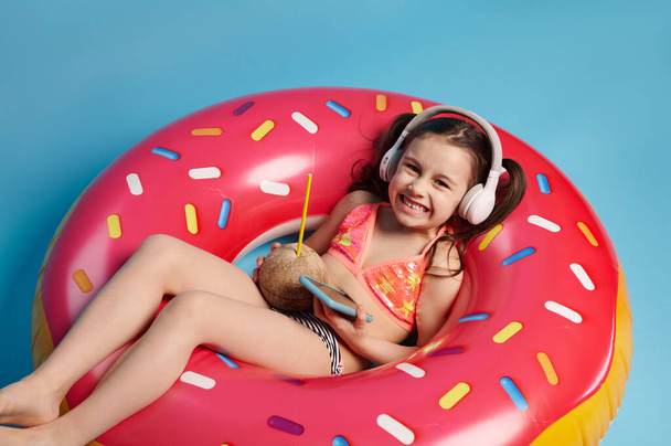 Menina pequena feliz com bebida de coco e smartphone moderno, ouve música em fones de ouvido sem fio e sorri olhando para a câmera, posando em um anel de natação inflável rosa no fundo de cor azul - Foto, Imagem