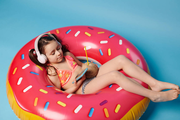 Ευτυχισμένο κοριτσάκι με μαγιό και ασύρματα ακουστικά, χαλαρώνοντας σε ένα φουσκωτό ροζ δαχτυλίδι κολυμπώντας ντόνατ, πίνοντας δροσιστικό νερό καρύδας, απολαμβάνοντας νέα εφαρμογή για κινητά στο smartphone, μπλε φόντο - Φωτογραφία, εικόνα