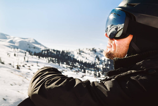 Close up of the ski goggles of caucasian man with the reflection of snowed mountains and sunburst. A mountain range reflected in the ski mask. Portrait of man at the ski resort - Foto, Imagem