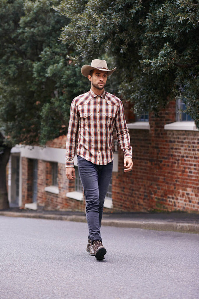 Miejski kowboj. przystojny mężczyzna ubrany w koszulę czekową i kowbojski kapelusz spacerujący wzdłuż drogi - Zdjęcie, obraz