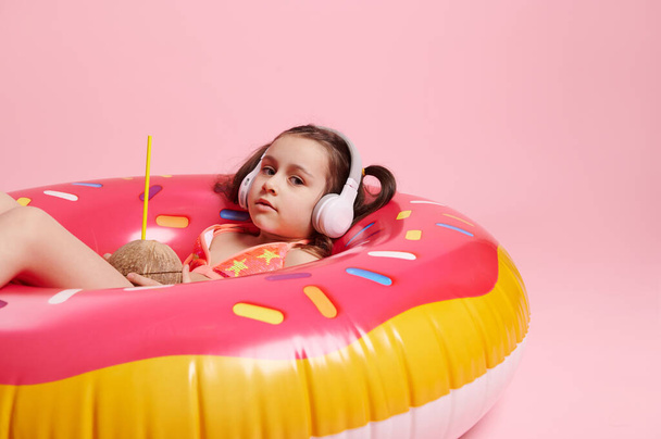 Κοριτσάκι με μαγιό, ακούγοντας μουσική σε ασύρματα ακουστικά, κρατώντας νερό κοκό ενώ βρίσκεται σε ροζ φουσκωτό σωλήνα κολύμβησης, απομονωμένο σε έγχρωμο φόντο. Καλοκαιρινές διακοπές και παραλία έννοια - Φωτογραφία, εικόνα
