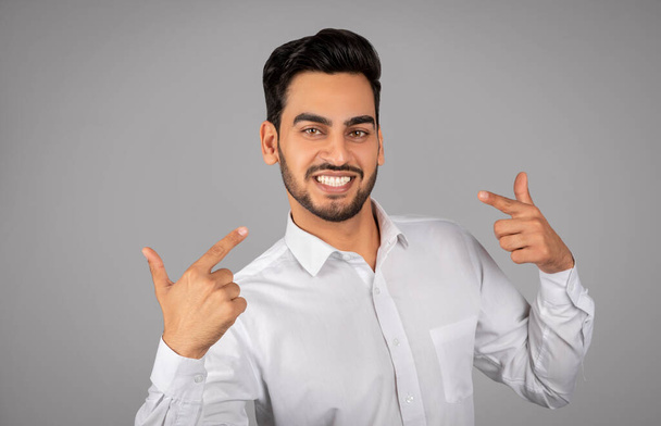 Mosolygó arab üzletember ujjal mutogat magára, és nézi a kamera, jóképű önbizalom közel-keleti férfi vállalkozó áll elszigetelt felett szürke stúdió háttér, másolás tér - Fotó, kép