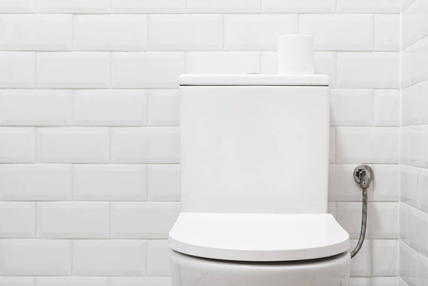 front view on clean wc bowl with closed lid and toilet paper on water tank on white brick tile background, restroom with light interior in modern apartment - Photo, Image