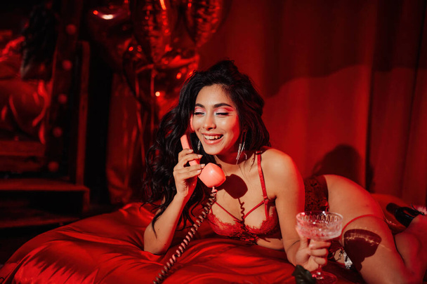 Asian model with long wavy hair posing with champagne on bed with red silk sheet, talking on retro phone on Valentine's Day - Zdjęcie, obraz