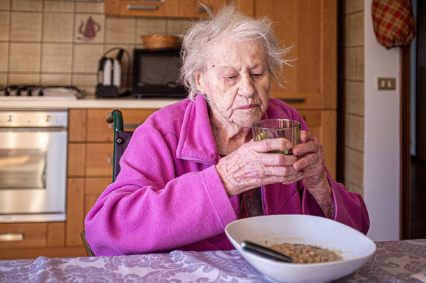 Old lady in a wheelchair with a glass of wine in hand eating alone in her kitchen - Foto, afbeelding