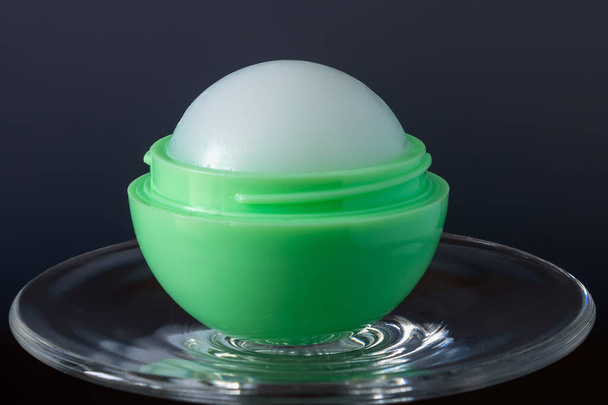 Female lip balm in green round ball on glass on dark background close up - Photo, image