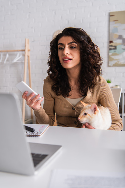 Copywriter holding cellphone and oriental cat near blurred laptop at home  - Фото, изображение