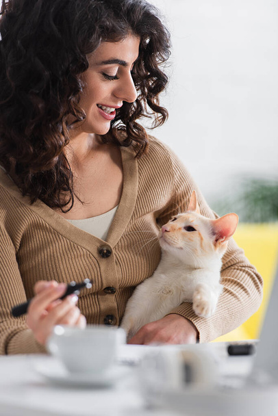 Mosolygós szövegíró holding marker és nézi keleti macska közel csésze és laptop otthon  - Fotó, kép
