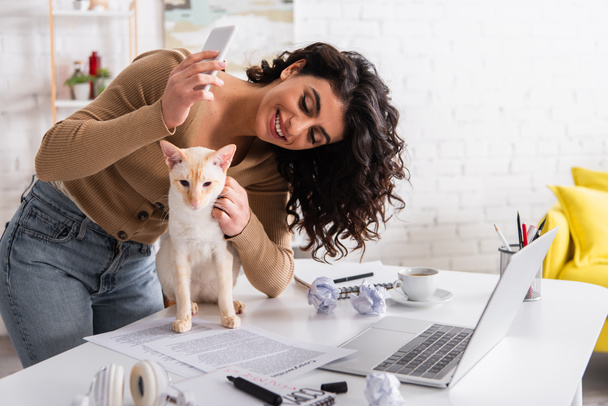 Cheerful copywriter holding smartphone and petting oriental cat near papers and laptop - Foto, immagini