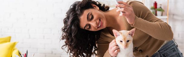 Usmívající se žena drží mobilní telefon a mazlit orientální kočka doma, prapor  - Fotografie, Obrázek