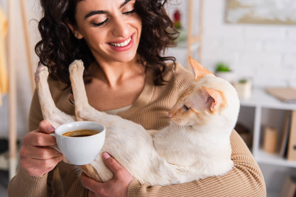 Smiling woman holding cup of coffee and oriental cat at home  - Фото, изображение