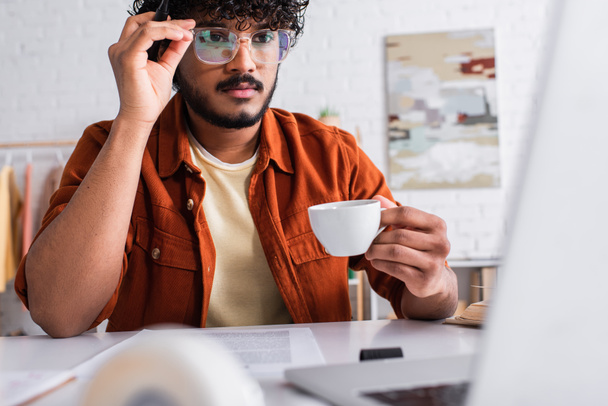 Ινδός copywriter σε γυαλιά κρατώντας φλιτζάνι καφέ κοντά θολή φορητό υπολογιστή στο σπίτι  - Φωτογραφία, εικόνα