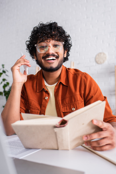 Cheerful indian copywriter in eyeglasses holding cellphone and book near papers at home  - 写真・画像