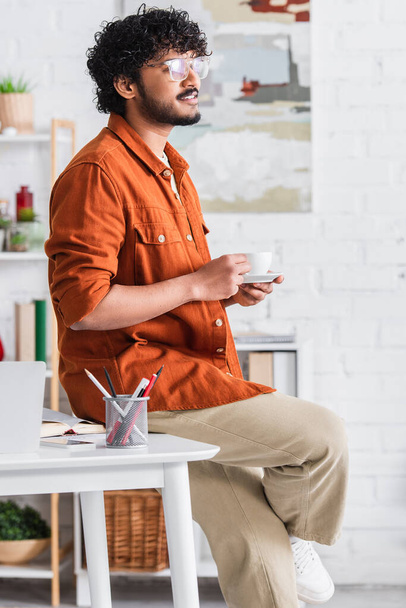 Smiling indian copywriter holding coffee near devices and book at home  - Fotó, kép
