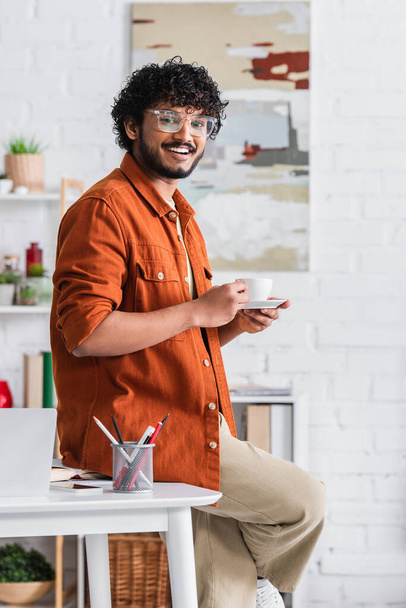Wesoły indyjski freelancer trzyma filiżankę kawy w pobliżu gadżetów w domu  - Zdjęcie, obraz