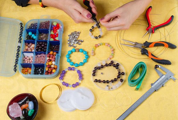 Schmuckherstellung. Herstellung eines Armbands aus bunten Perlen. Weibliche Hände mit einem Werkzeug auf gelbem Hintergrund.  - Foto, Bild
