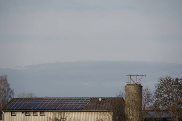 Moduły słoneczne na dachu dużej stodoły - farma producentów energii elektrycznej. Słońce świeci jasno na dachu i pozwala modułom świecić w świetle: niebo powyżej jest zachmurzone - Zdjęcie, obraz