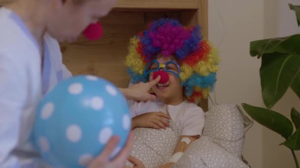Medical sister in a professional uniform with a red nose plays with a boy in his hospital ward. High quality 4k footage - Záběry, video