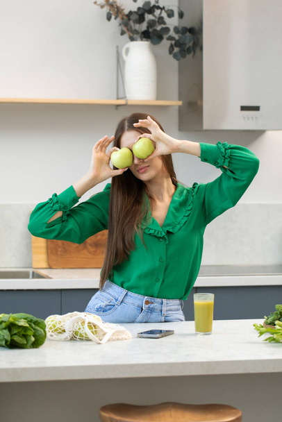 An attractive woman closes her eyes with green apples and smiles while standing in the kitchen. The concept of healthy eating, vegetarianism, detox. - Fotografie, Obrázek