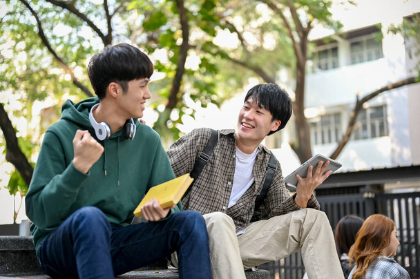 Fröhlicher junger asiatischer männlicher College-Student genießt das Gespräch mit seinem Freund während der Pause im Universitätspark. - Foto, Bild