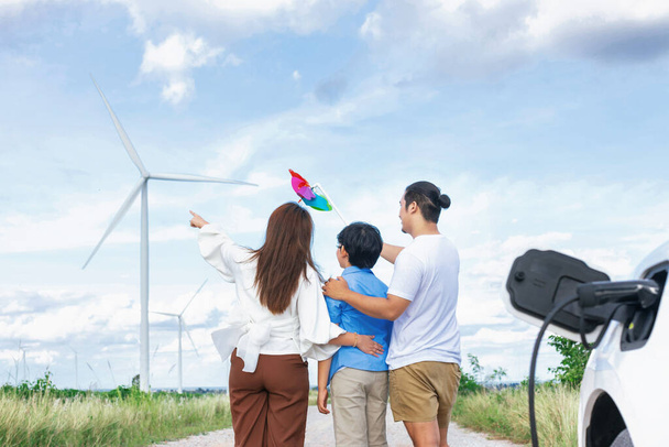 Concept of progressive happy family holding windmill toy and relax at wind farm with electric vehicle. Electric vehicle driven by clean renewable energy from wind turbine generator to charger station. - Foto, Bild