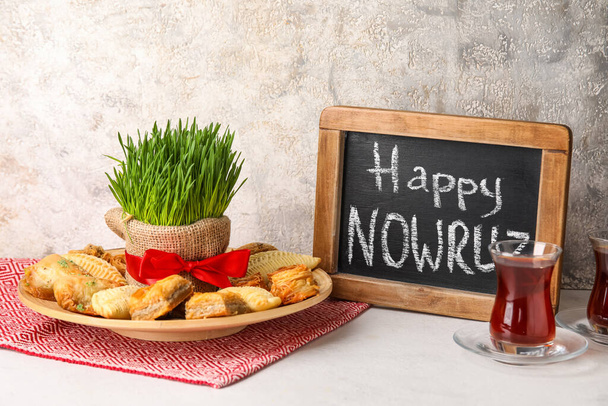 Крейдяна дошка з текстом HAPPY NOWRUZ, ласощі, трава і склянка чаю на столі біля гранжевої стіни
 - Фото, зображення