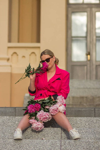 Smiling successful businesswoman in crimson dress and glasses holding bouquet of pink peonies. Happy young blonde with bunch of fresh flowers. St. Valentines Day, International Womens Day concept. - Фото, зображення