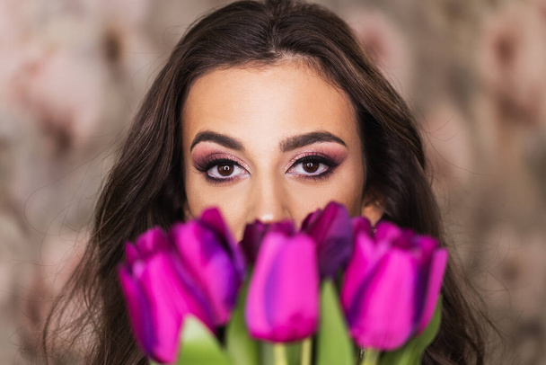 Muotokuva viehättävästä nuoresta naisesta, jolla on kauniit silmät, joka pitää kukkakimpun violetteja tulppaaneja ja edustaa mukavaa meikkiä kasvoillaan.. - Valokuva, kuva