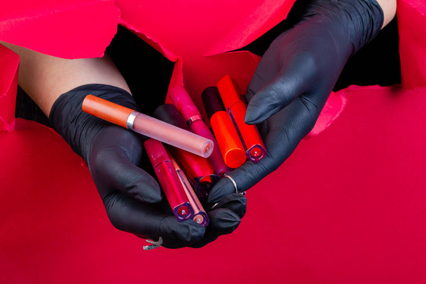 Gloved hands holding pile of various lip gloss sticks. Breaking through a red paper wall. - Foto, afbeelding