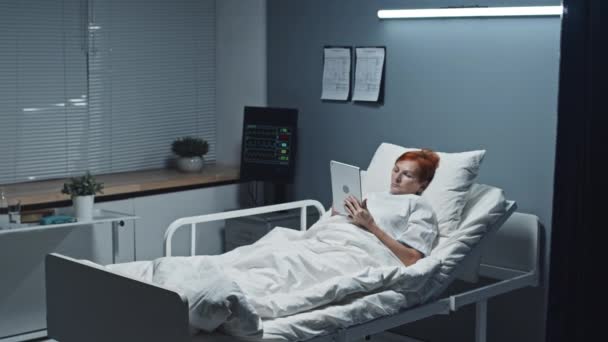 Seguimiento de la mujer caucásica madura de pelo corto acostada en la cama del hospital, usando tableta, recuperándose después de la cirugía - Metraje, vídeo