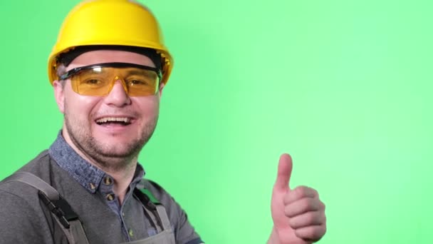 Happy smiling worker showing thumb up on green background in studio. Advertising shooting of a young builder with a European appearance. - 映像、動画