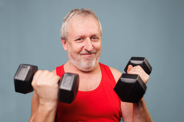 Athletic senior citizen with a twist This studio shot features a 60s male pensioner lifting dumbbells with an energetic and athletic posture. But the comical expression on his face - Foto, Imagen