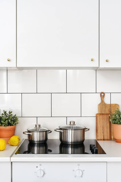 λεπτομέρεια στην κουζίνα με φως εσωτερικό, λευκό ντουλάπι έπιπλα, πλακάκι στον τοίχο, σύγχρονες ηλεκτρικές εστίες, ανοξείδωτο χάλυβα κατσαρόλα με καπάκι και φυτό σε κατσαρόλα - Φωτογραφία, εικόνα