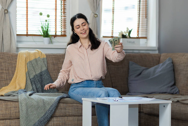 Purchase of real estate. Happy young caucasian woman sitting on sofa puts money on table. Concept of leasing, business and mortgage. - Photo, image