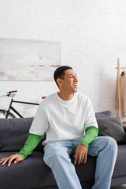 onnellinen afrikkalainen amerikkalainen mies katselee pois, kun istuu sohvalla olohuoneessa - Valokuva, kuva