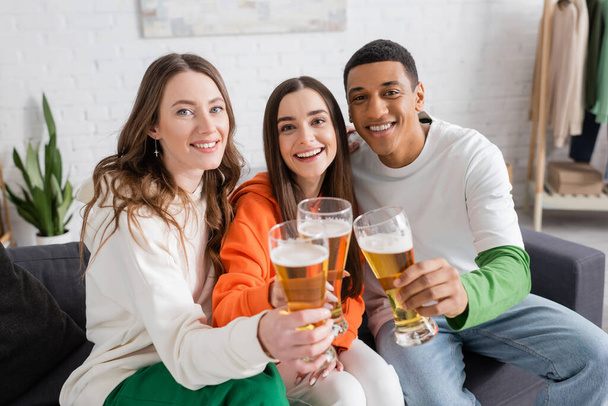 cheerful women and african american man clinking glasses of beer while looking at camera in living room  - Photo, Image