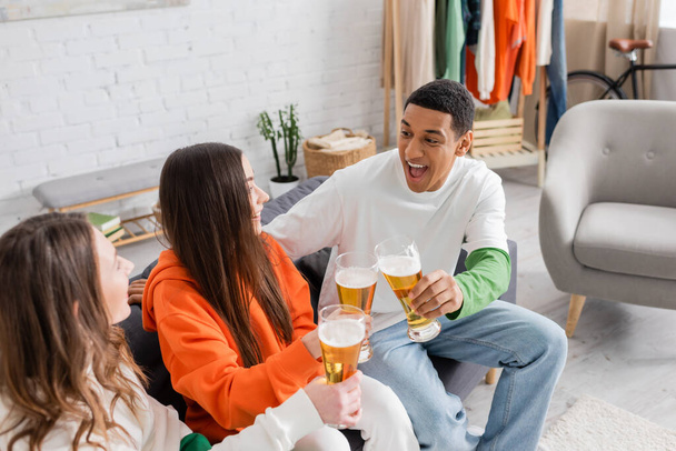 cheerful women and amazed african american man clinking glasses of beer in living room  - Foto, immagini