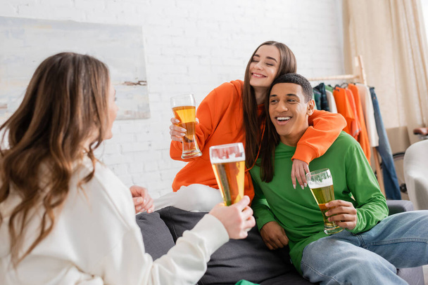 smiling woman hugging cheerful african american man while holding glass of beer and looking at friend  - Fotografie, Obrázek
