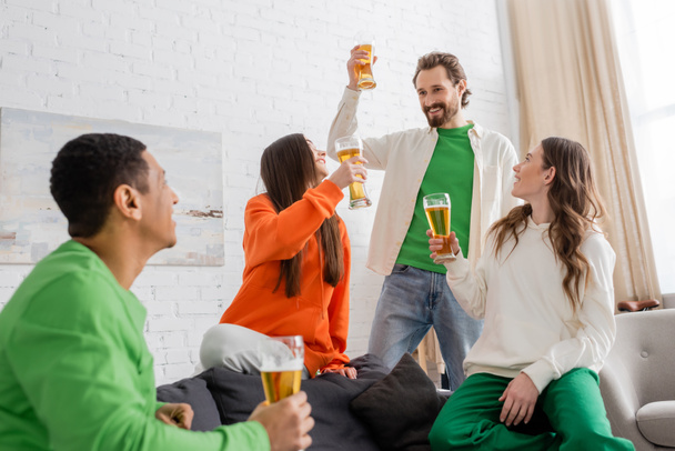 felice uomo barbuto brindare vetro di birra vicino alle donne e guardando amico afro-americano in soggiorno  - Foto, immagini