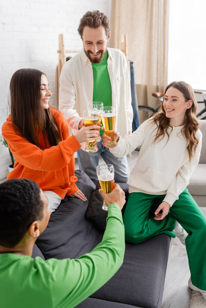 happy multiethnic friends toasting glasses of beer in living room  - Fotografie, Obrázek