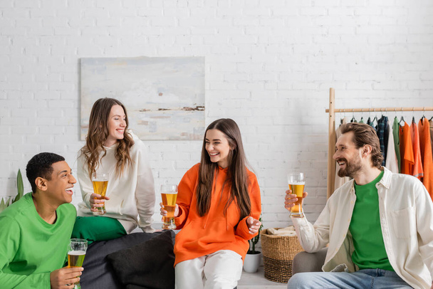 cheerful multiethnic friends holding glasses of beer while smiling in living room  - Photo, image