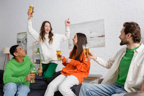 excited woman with glass of beer gesturing near interracial friends in living room  - Foto, immagini
