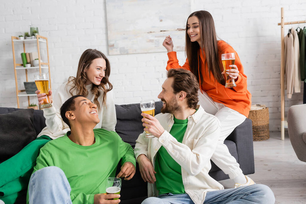happy interracial group of friends holding glasses of beer while looking at each other in living room  - Фото, изображение