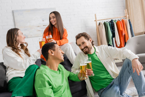 cheerful multicultural men and women clinking glasses of beer while chatting in living room  - Foto, Bild