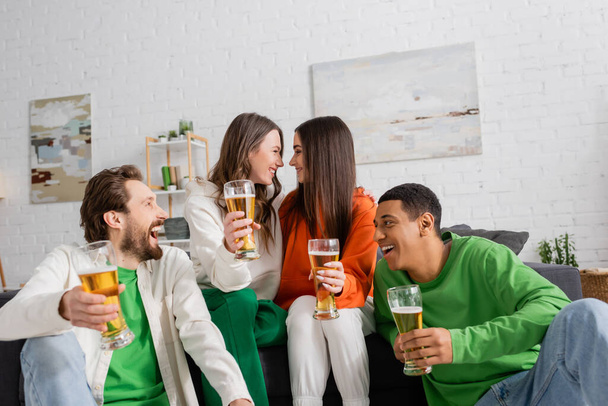 positive and interracial group of friends holding glasses of beer while looking at each other in living room - Foto, imagen
