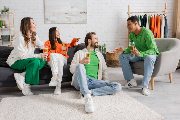 cheerful woman sitting near friend and pointing at interracial men talking in living room  - Foto, Bild