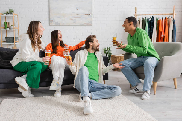 cheerful multicultural men and women holding glasses of beer while chatting in living room  - Foto, Bild