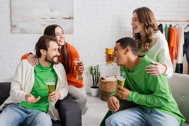 happy women hugging interracial men while holding glasses of beer in living room  - Foto, immagini